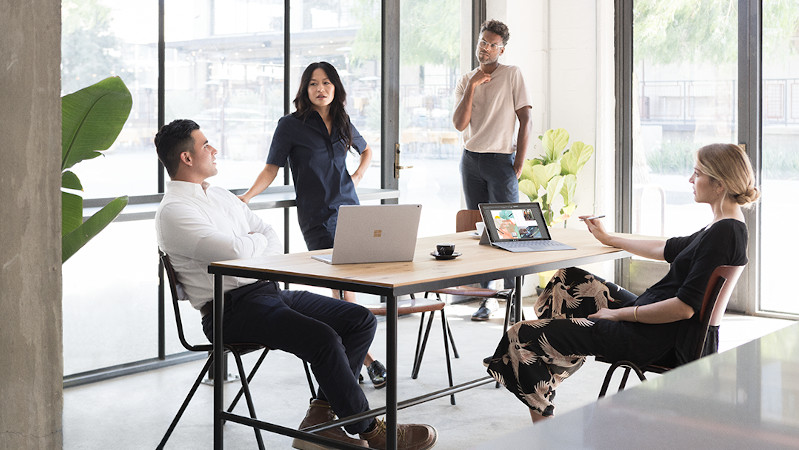 Zwei Frauen und zwei M&auml;nner sitzen bzw. stehen an einem Tisch in einem Besprechungsraum mit Surface-Ger&auml;ten vor ihnen