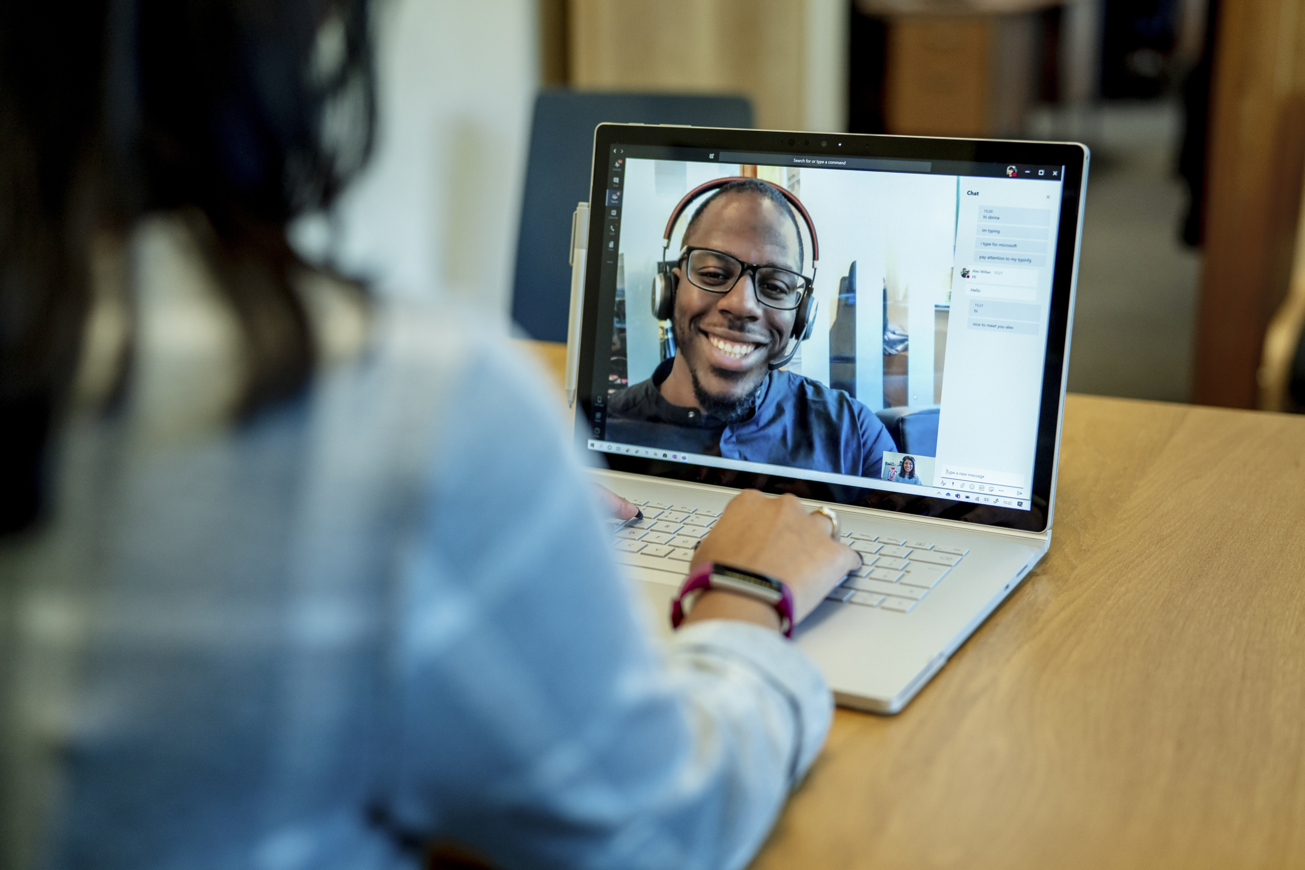 Woman at a desk using a Surface laptop to make a Microsoft Teams video call with one man smiling and wearing a headset. Business Voice conference call/meeting device is in the background