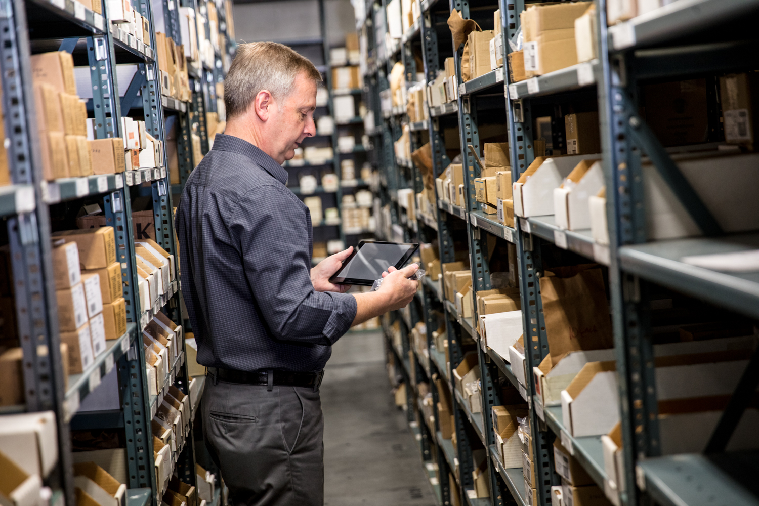 Male employee in warehouse storage room holding a tablet in one hand and small item in the other. He is standing between metal warehouse racks filled with pacakges