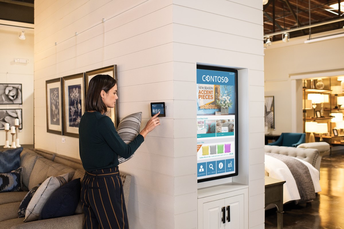 Female employee standing on home furnishings sales floor in commercial retail store, holding a pillow and using a wall tablet