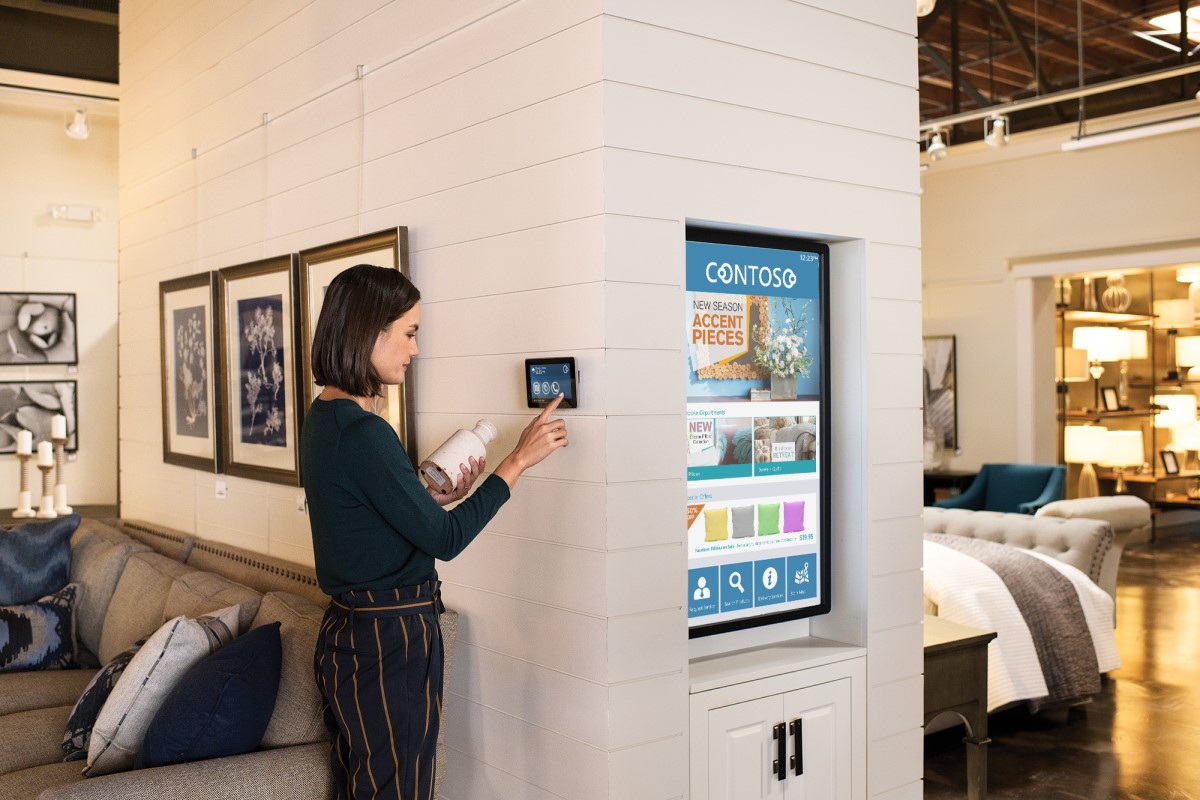 Female employee standing on home furnishings sales floor in commercial retail store, holding a vase and using a wall tablet