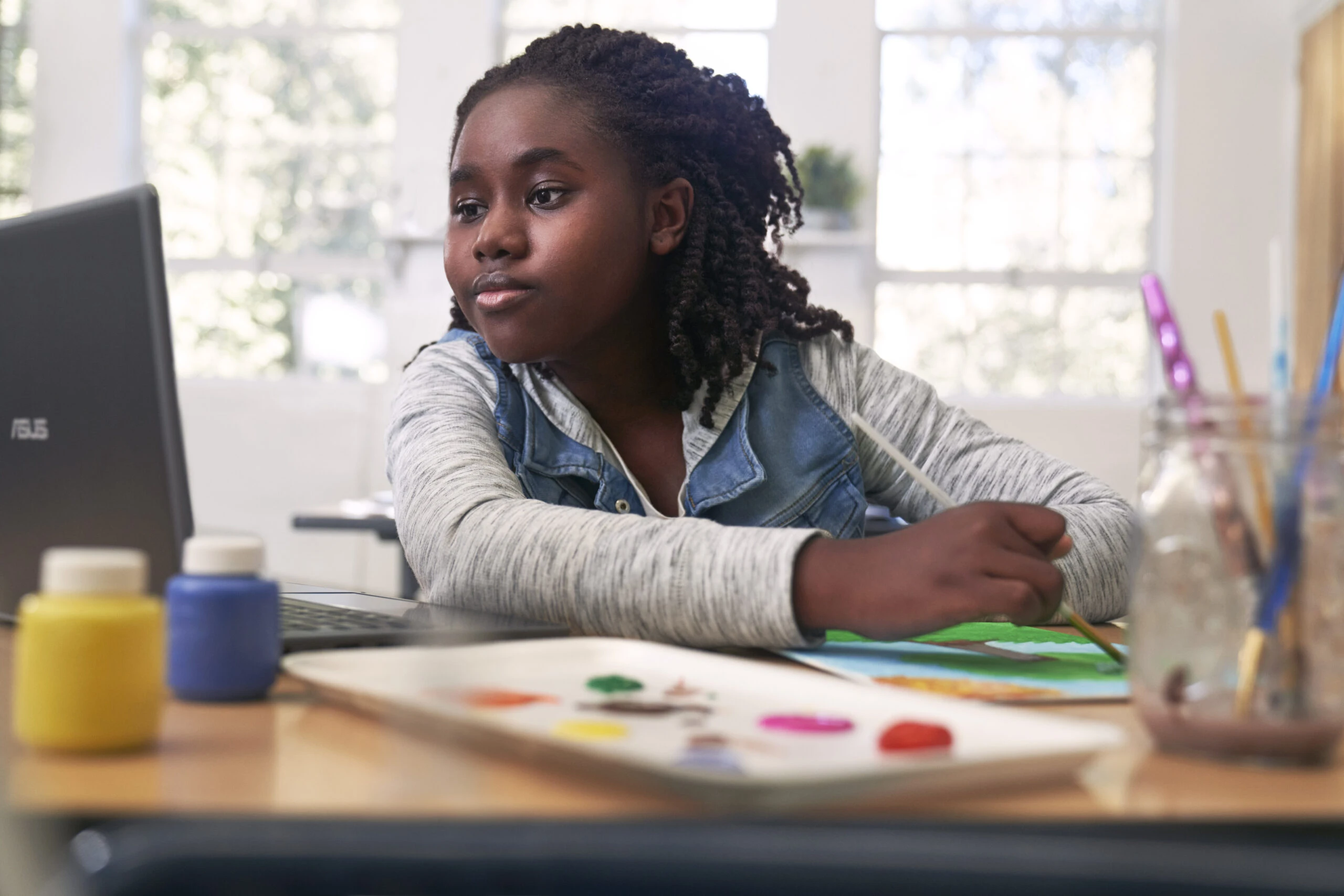 Female K-12 student sitting at a desk painting a tree while looking at a laptop