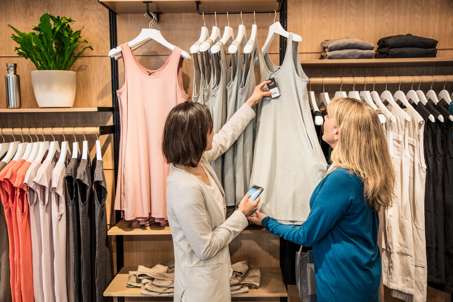 Photo of a female store employee using phone standing next to female consumer in front of retail clothing display. The employee is checking the price tag