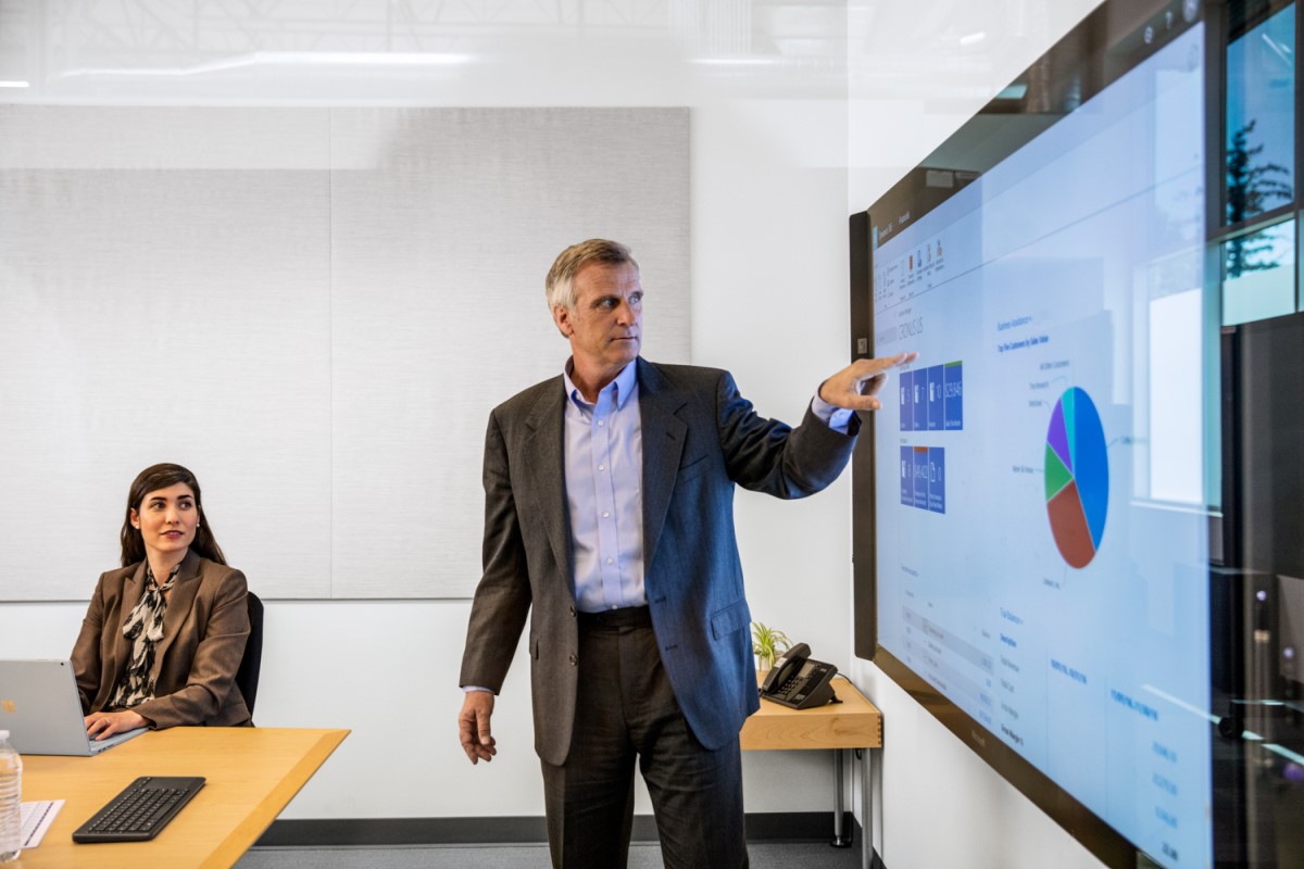Male businessman in suit giving presentation in office conference room. He is pointing at a large monitor screen, which displays a pie chart and several statistics/monetary values