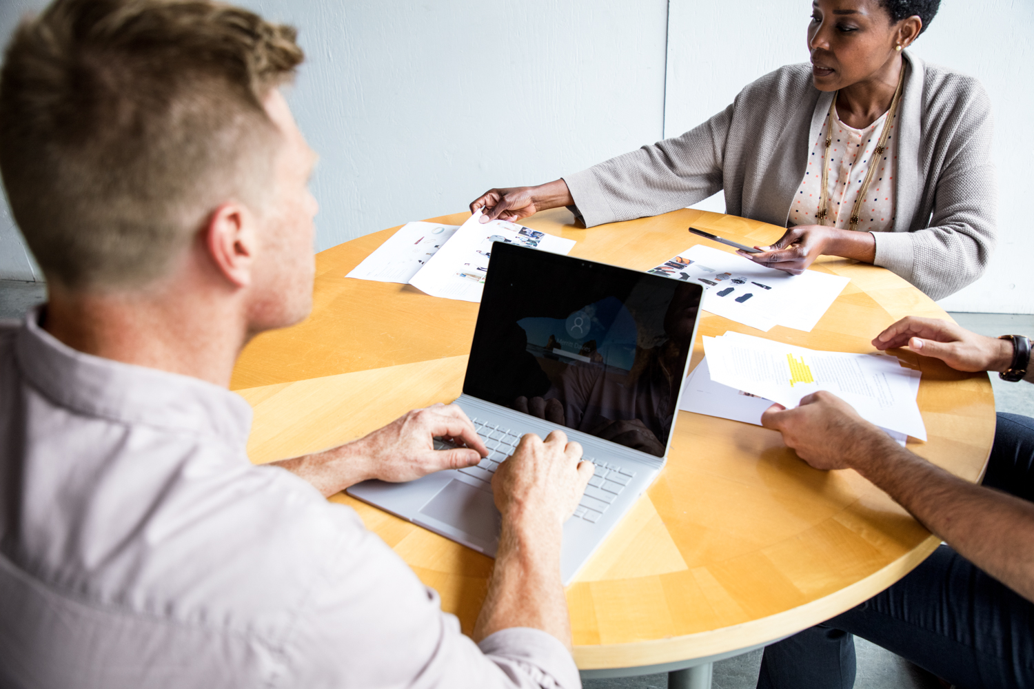 Female and male workers sitting at round table in discussion. A third persons (male) hands are also visible. The man is using a Surface Laptop, while the woman is reviewing paperwork