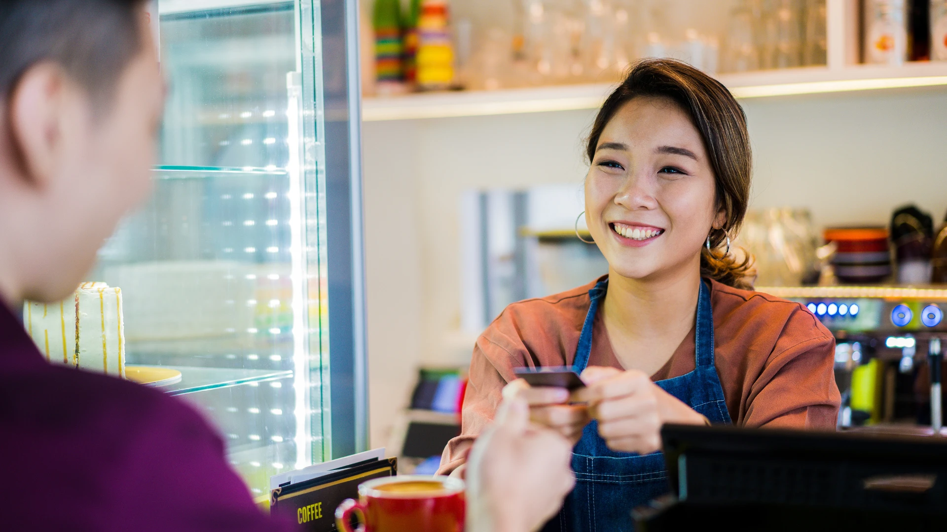 Person at a café counter holding a payment card during a customer transaction, with a coffee machine and display case visible in the background