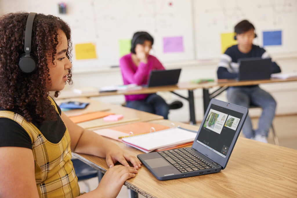 A student sitting at a desk in a classroom wearing headphones and working on an assignment on a laptop. Two other students are working on laptops in the background.