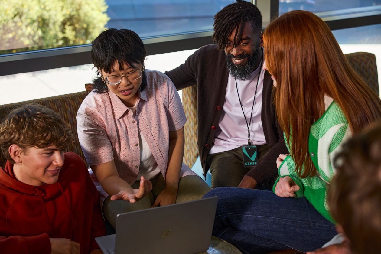 An instructor and a group of students collaborate on a laptop in a common area at a college