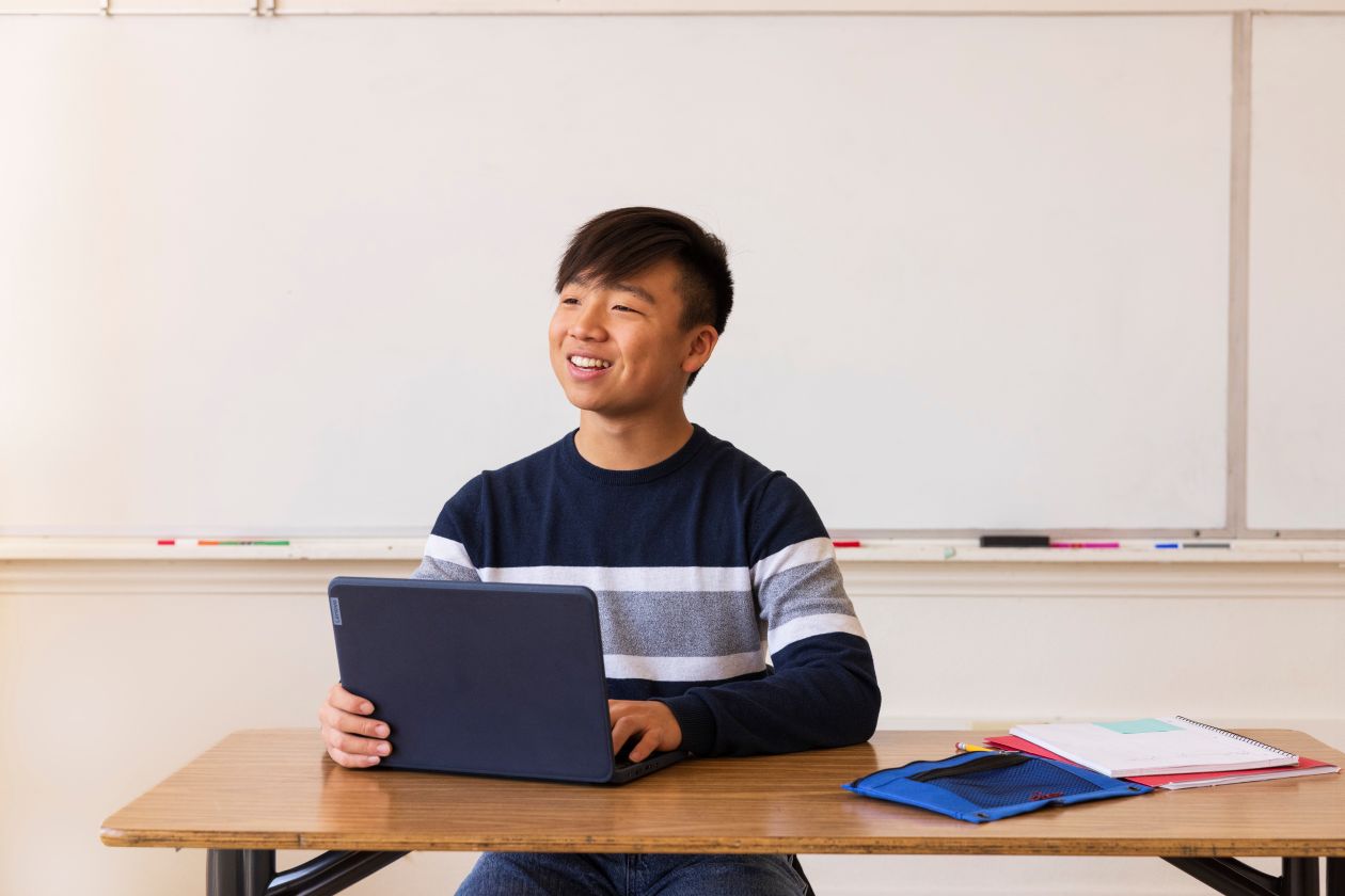 An older student sitting at a table in a school classroom while using a laptop