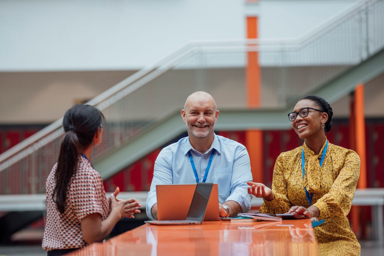 Three educators with laptops sit at a table and collaborate in a school common area