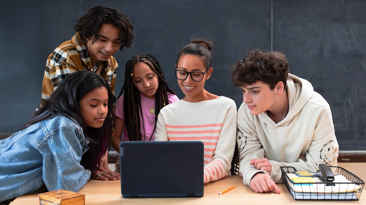 An educator and four students looking at a laptop at the educator’s desk in a school classroom