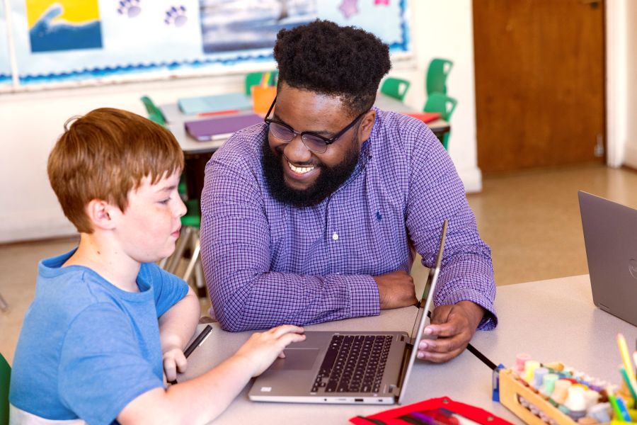 An educator kneeling at a student’s desk and helping him with schoolwork on his laptop.