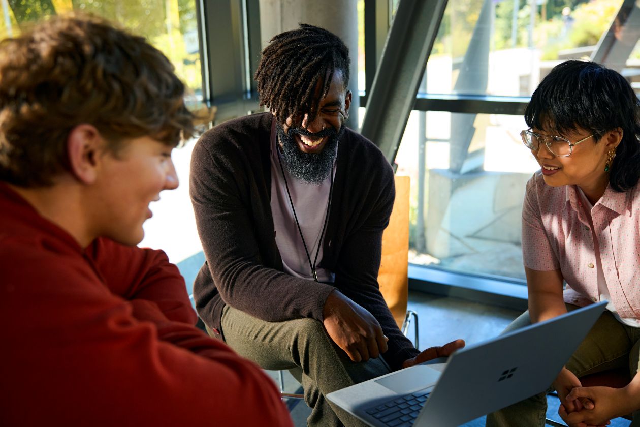 An instructor and a group of students collaborate on a laptop in a common area at a college