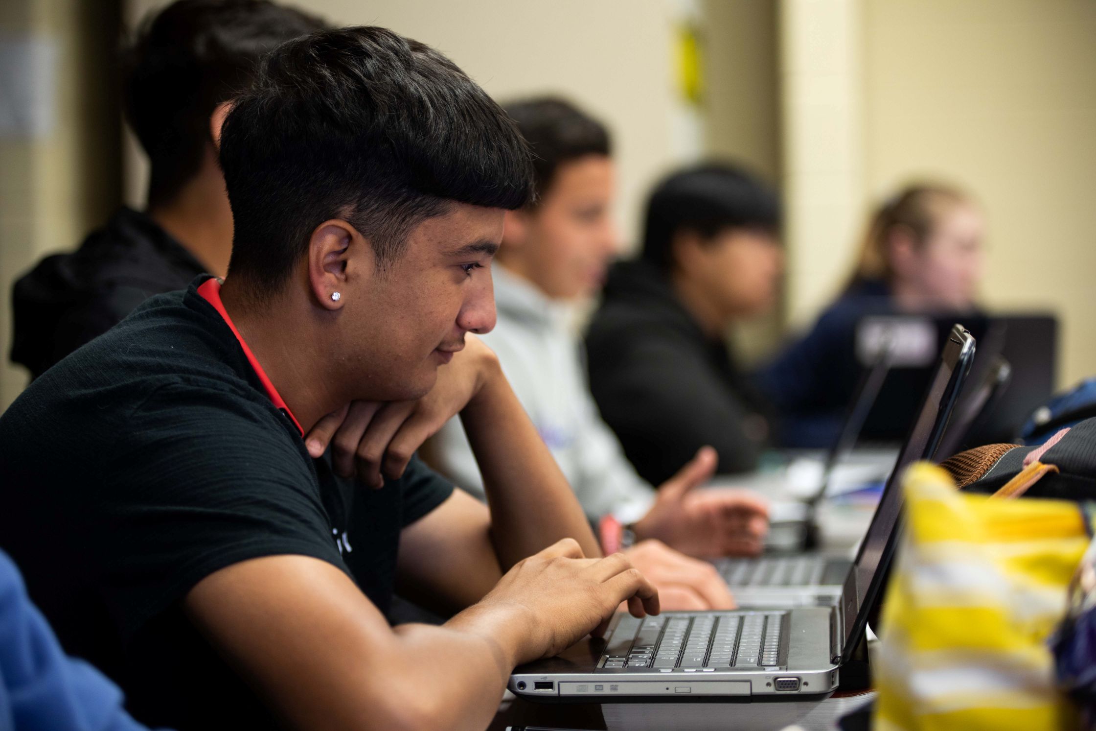 A classroom scene showing a row of students using laptops, focused on single student in the foreground