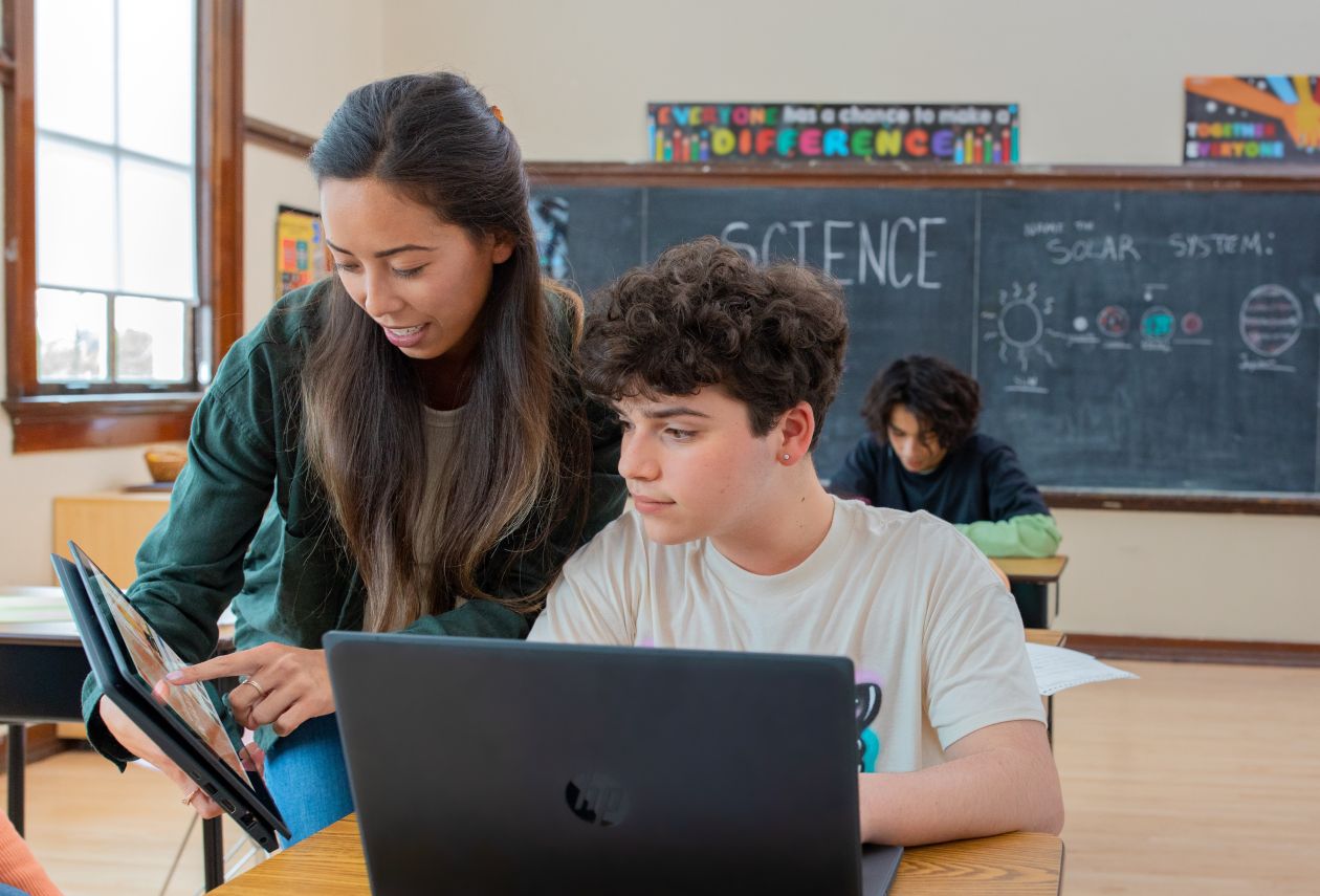 A teacher engages with a student at his desk. Theyre both using laptops