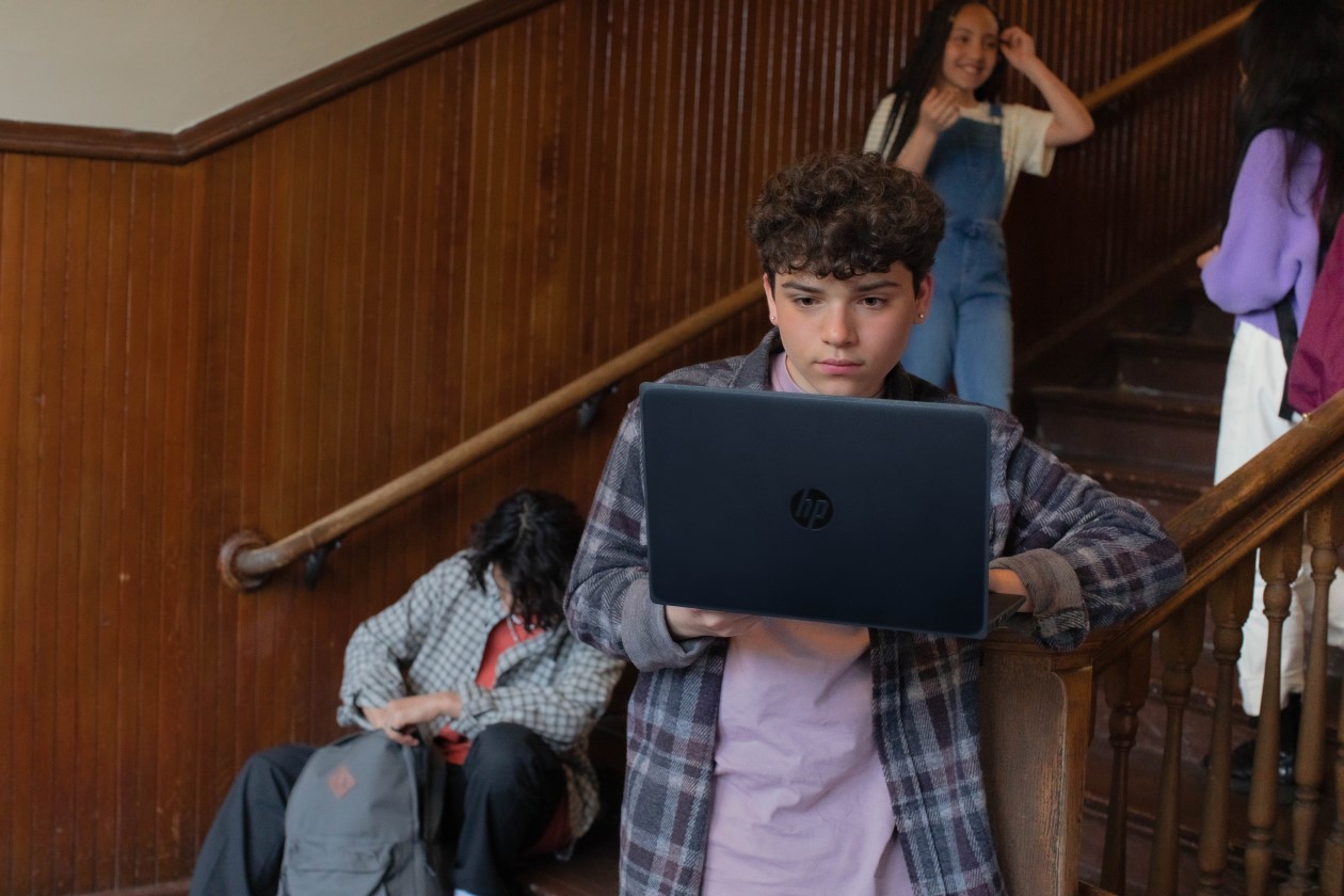 A male pre-teen student standing at the bottom of a staircase at school and working on a laptop. There are three other students hanging out on the stairs behind him