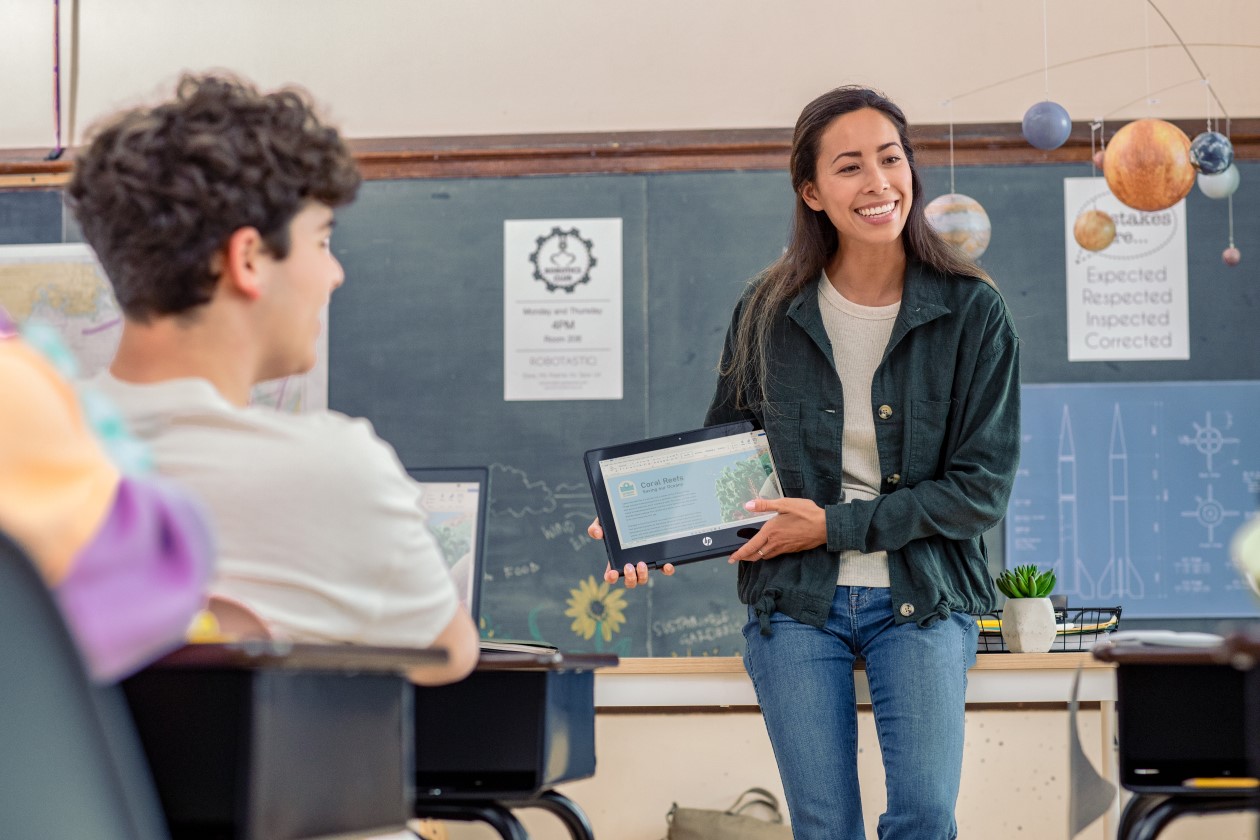 Decorative. A teacher holding a folded laptop in front of students in a classroom