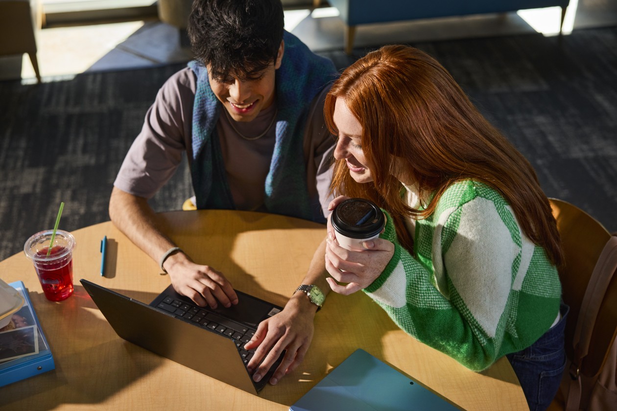 Two higher education students look at the screen of a laptop while sitting at a table and collaborating