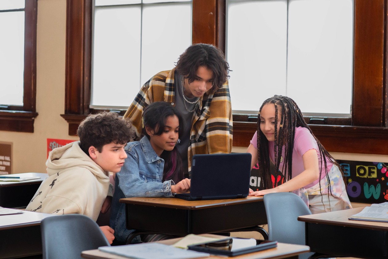 Four students gathered around an open laptop at a desk in a school classroom