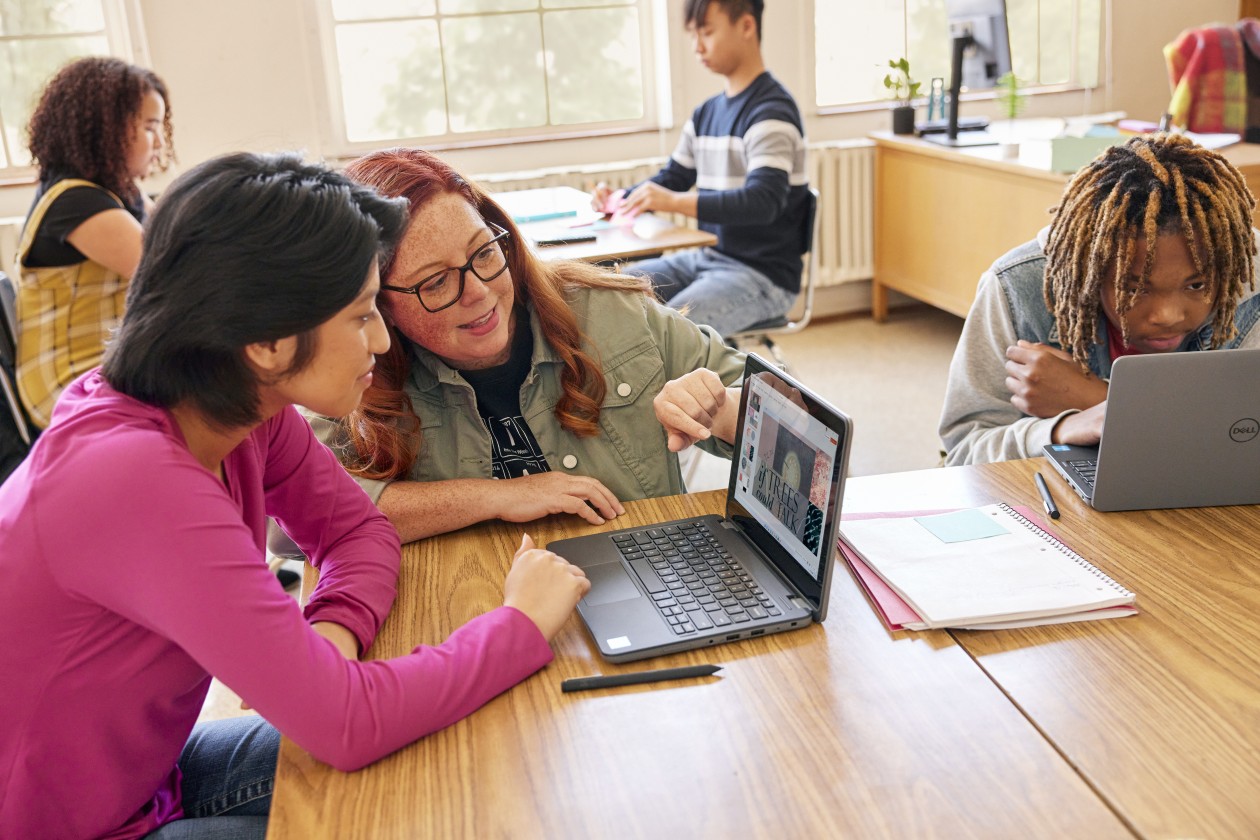 A classroom scene with students at communal desks and an educator leans in to help one of the students
