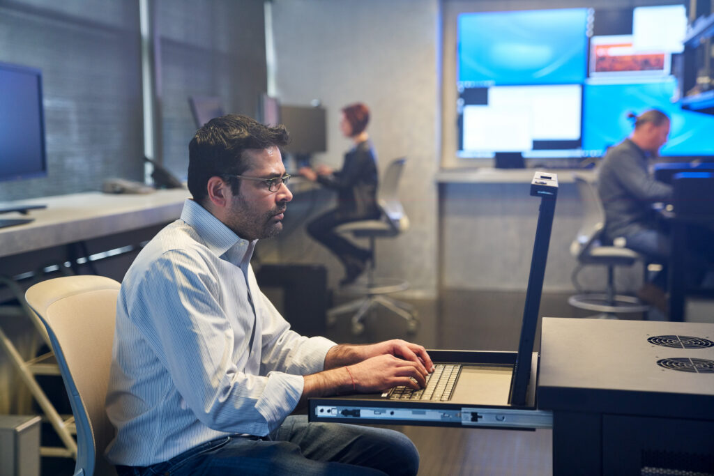 An IT professional works at a laptop in a security operations center, with colleagues and multiple monitoring screens visible in the background.