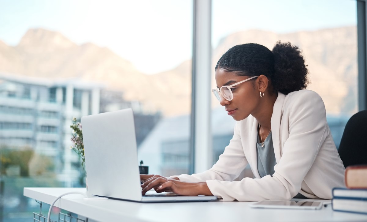 Picture of a woman wearing a white blazer and glasses, looking down at a laptop on her desk