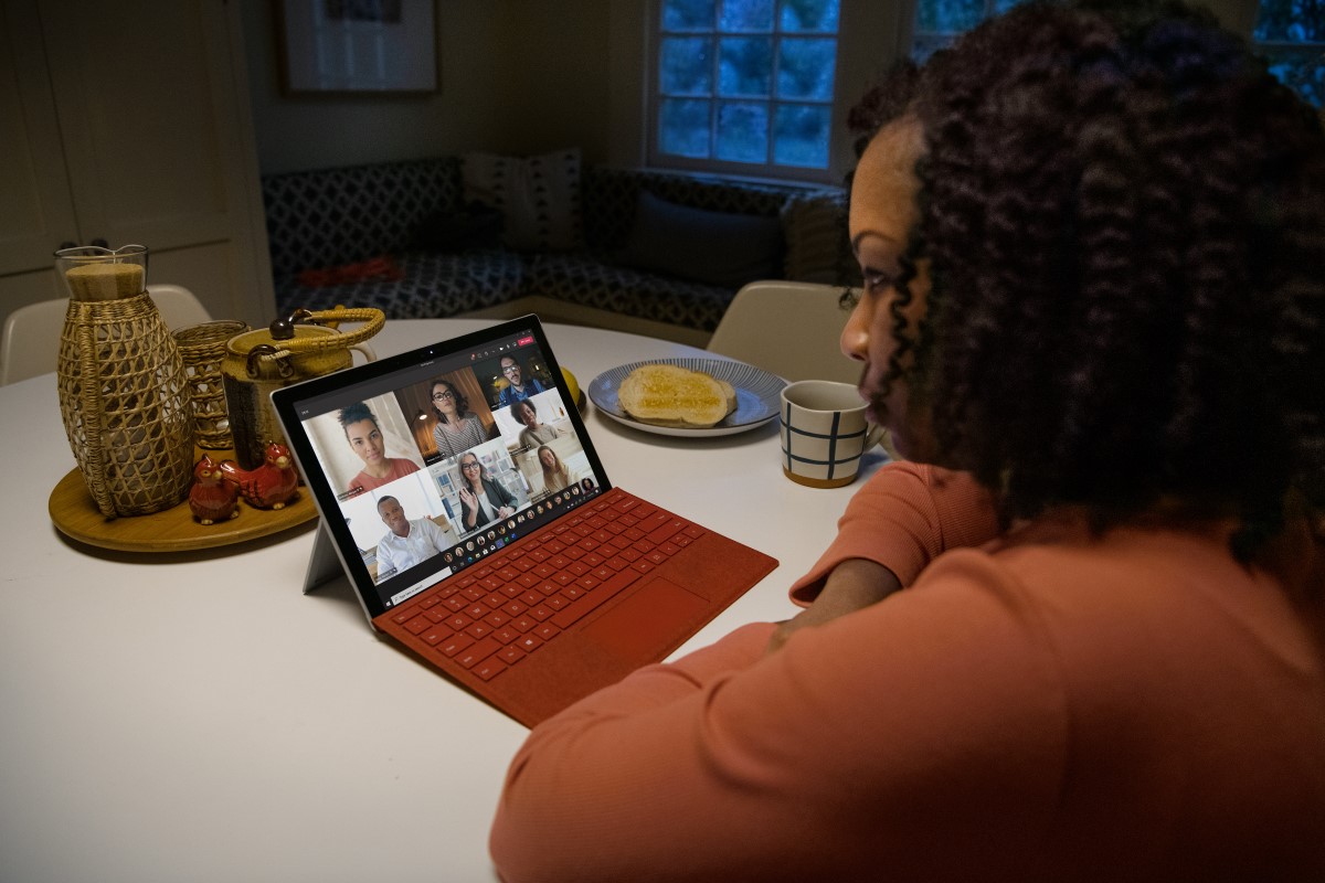 Over the shoulder photo of an adult female in kitchen participating in a Microsoft Teams video call on a Surface Pro with Poppy Red type cover