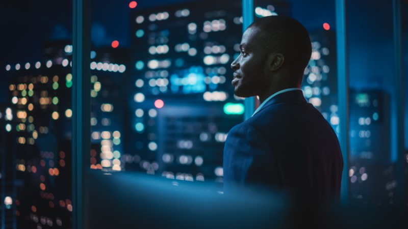 A businessman in the financial services industry looks out an office window, the lights of nearby buildings in the background, as he reflects on the new year ahead