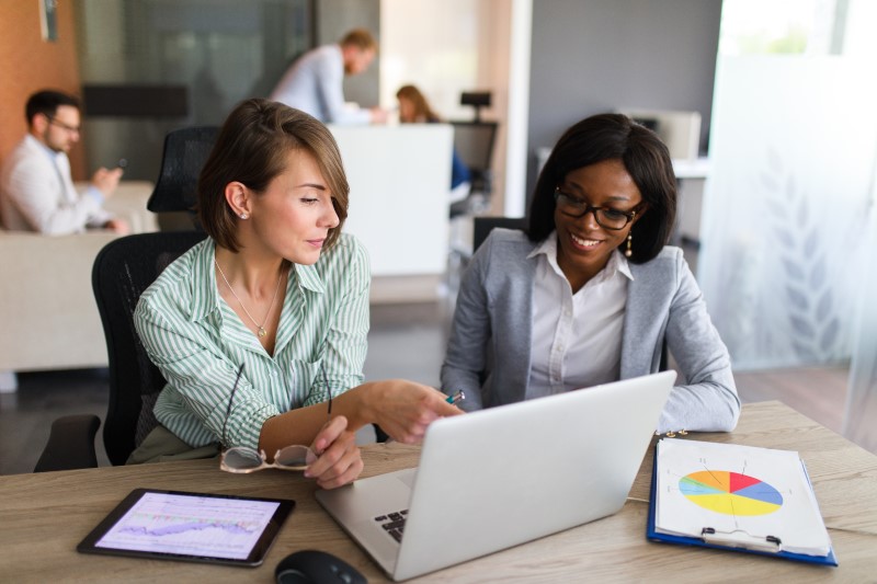 Two businesswomen are in the office using laptop, digital tablet and chart papers while working together
