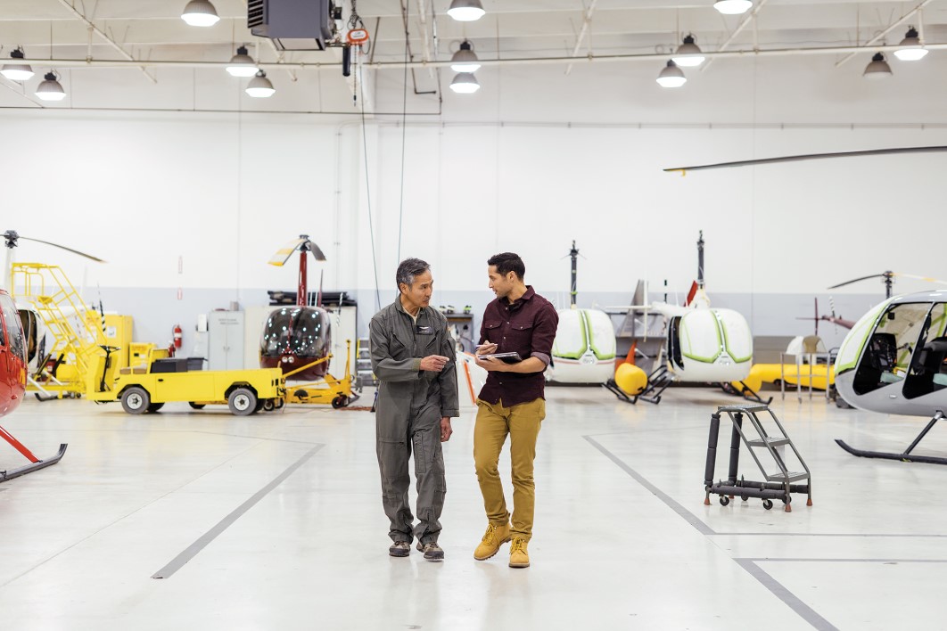 Two male first line workers in conversation while walking among assembled helicopters on factory floor in commercial manufacturing plant