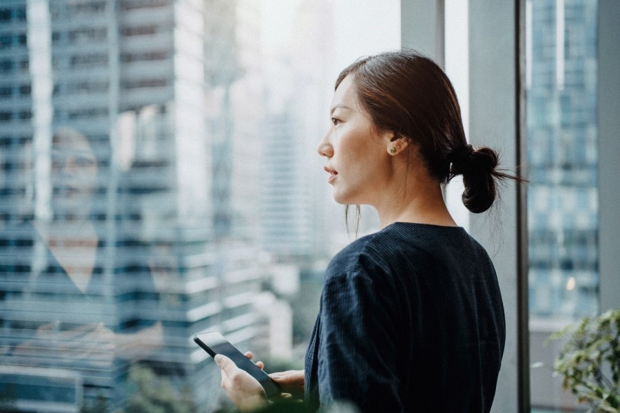 Young businesswoman holding a phone, looking out her office window overlooking the city