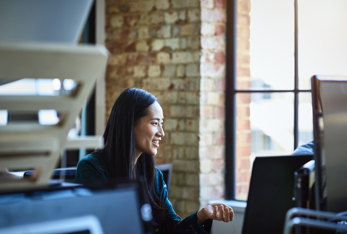 Office worker, smiling and looking away, in contemporary studio with bare brick walls, sitting in front of a desktop computer