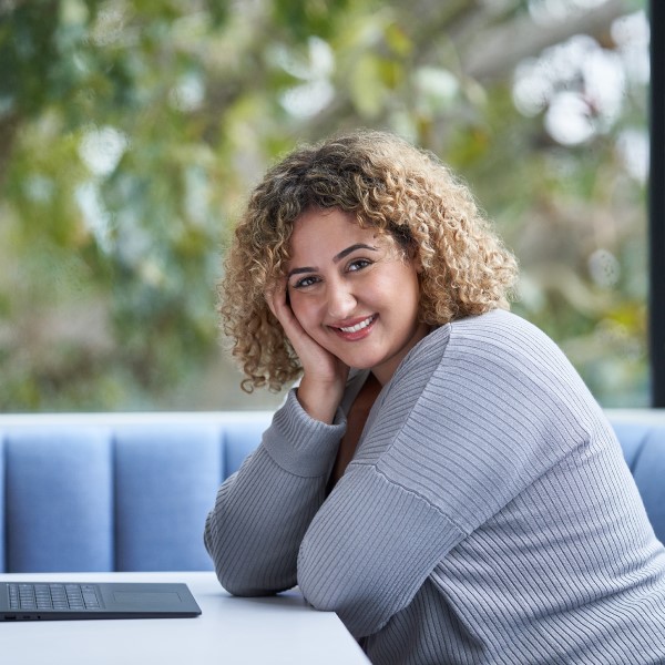 A person sitting at a table at their laptop, looking at the camera, smiling