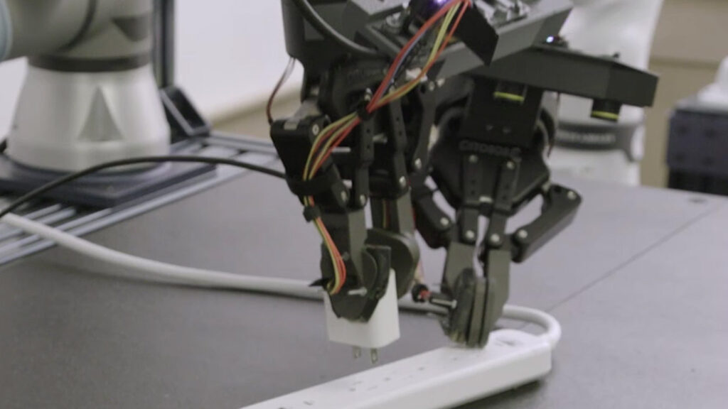 Rho-Alpha | close up image of a robotic arm and hand holding down a power outlet strip while unplugging a small charging adapter.