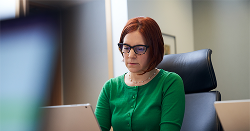 An image of a red-haired woman with a green shirt and glasses, sitting inside an office working on her laptop