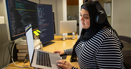 An image of a female developer coding at her desk, wearing hijab and headphones