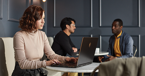 A remote female tech worker on her laptop while two co-workers have a discussion in the backgroud