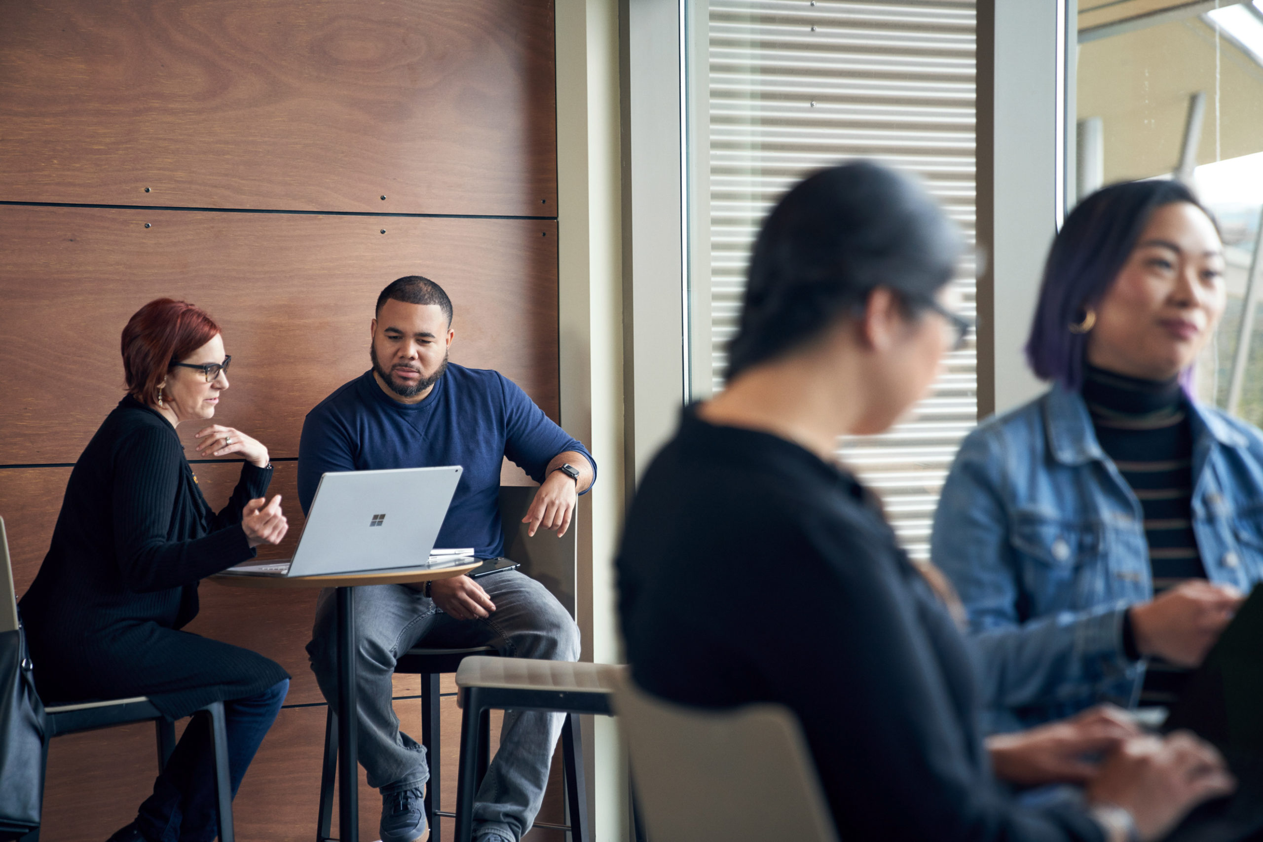Man and woman meeting together and reviewing content on a Surface laptop at a high table with two blurred women in the foreground