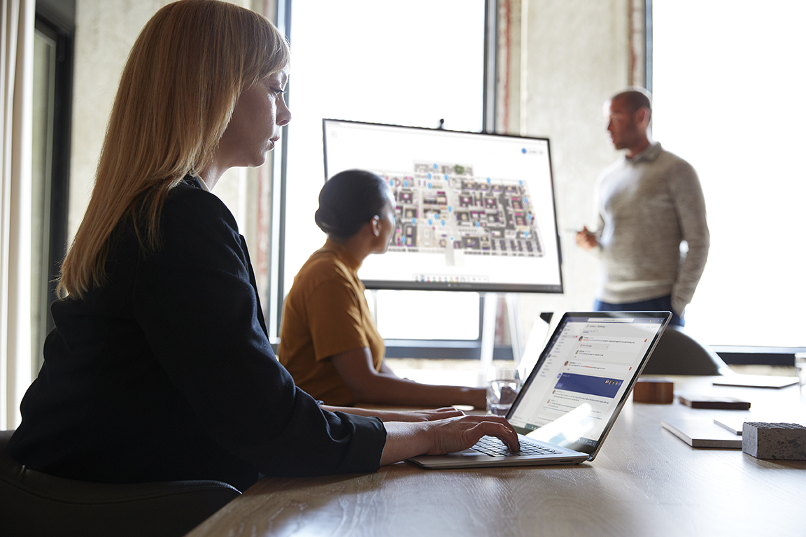 Three adults are collaborating in a conference room. One male is standing next to a Microsoft Surface Hub 50” with Microsoft Whiteboard screen shown