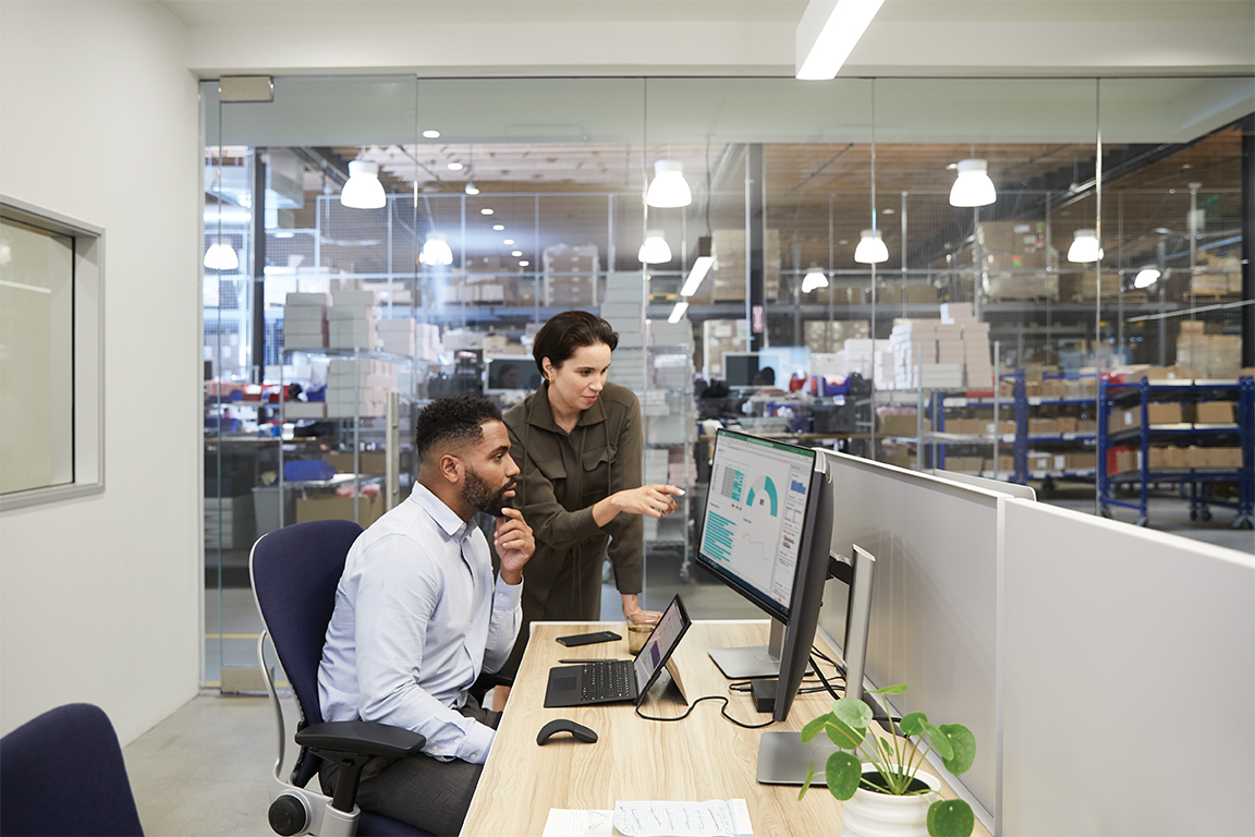 Two adults in an office adjacent to a warehouse setting collaborating while overlooking dual monitors