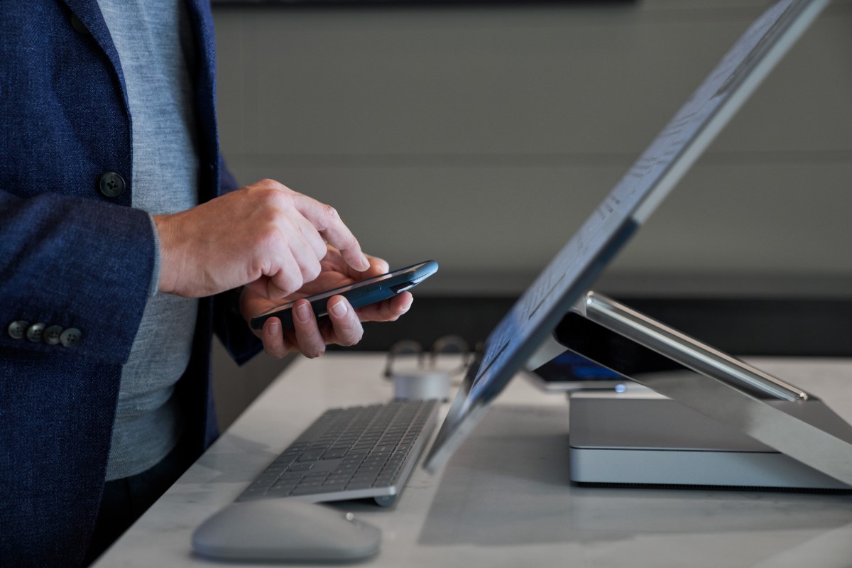 Side view close-up of a man typing on his phone while standing behind a Microsoft Surface Studio