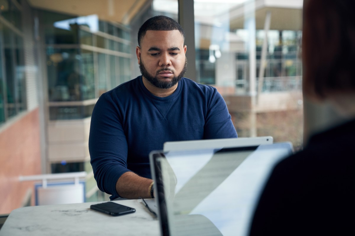 Man sitting at a community table in common area typing on a Surface laptop with a phone at his side