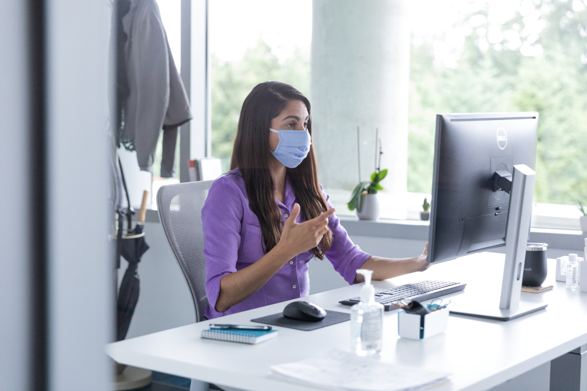 A female employee wearing a mask and talking on a video call with hand sanitizer on her desk, socially distancing from others