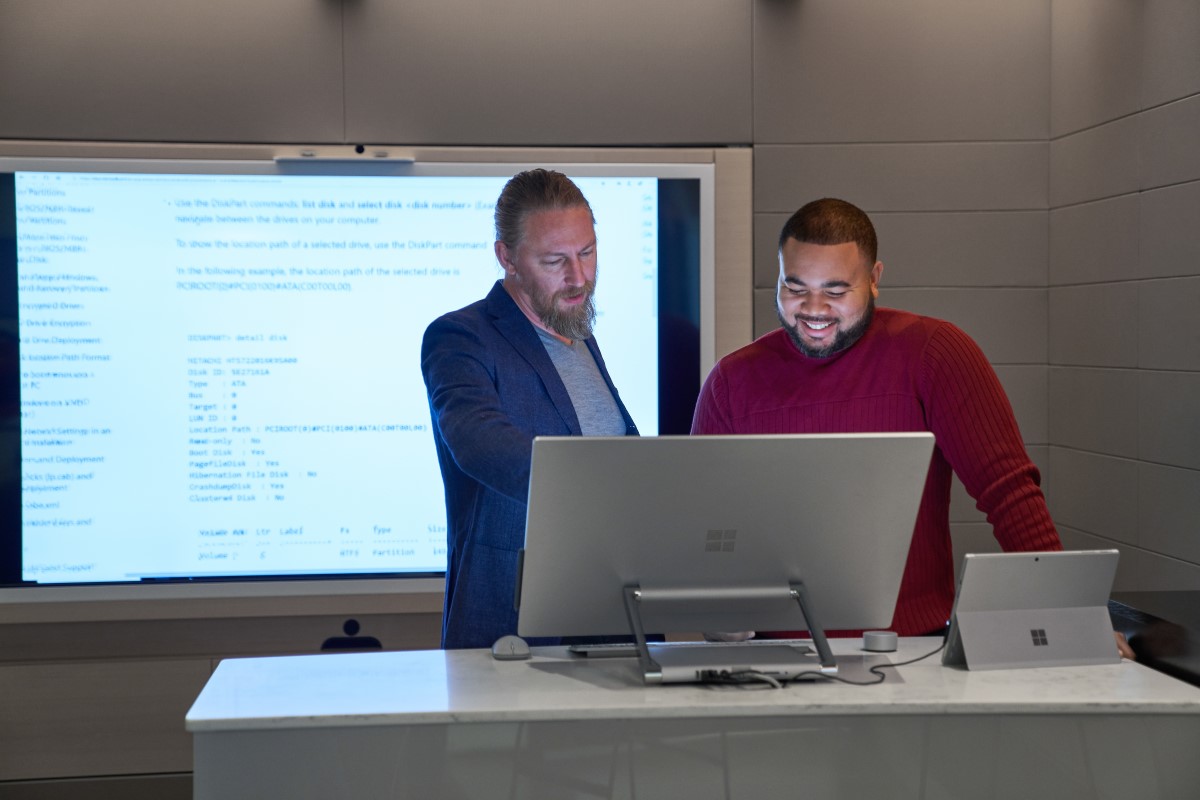 Forward facing view of two men working on a Microsoft Surface Studio with a larger blurred screen/display behind them