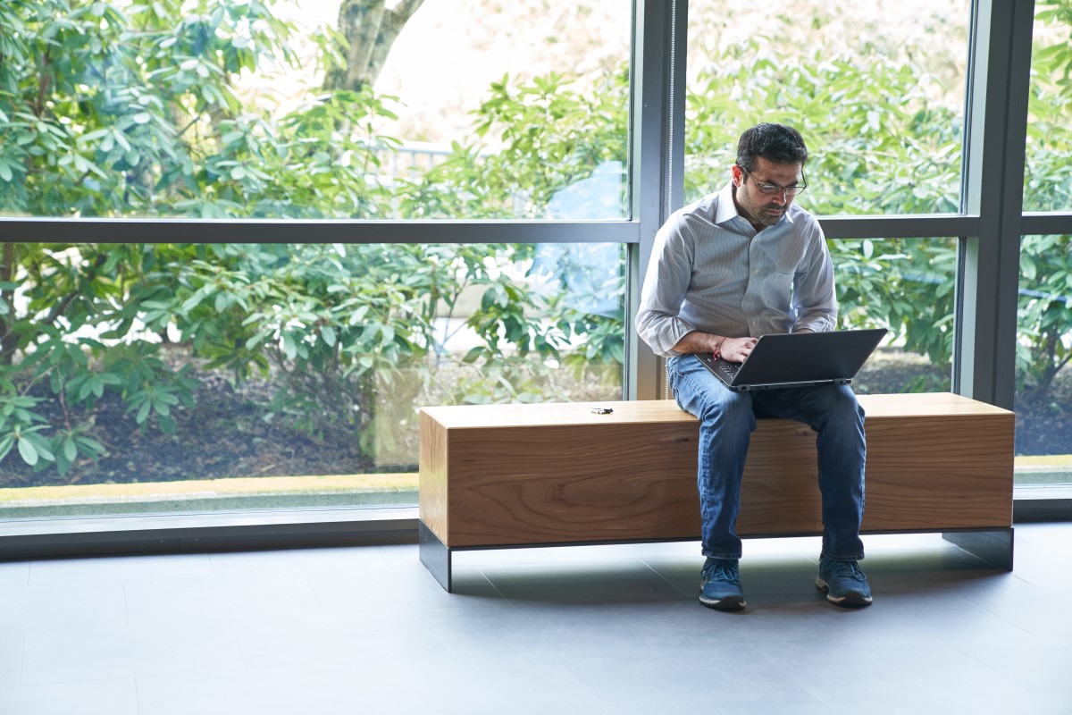 Man sitting on an indoor bench in front of windows working on a laptop with a security key fob or USB drive next to him on the bench