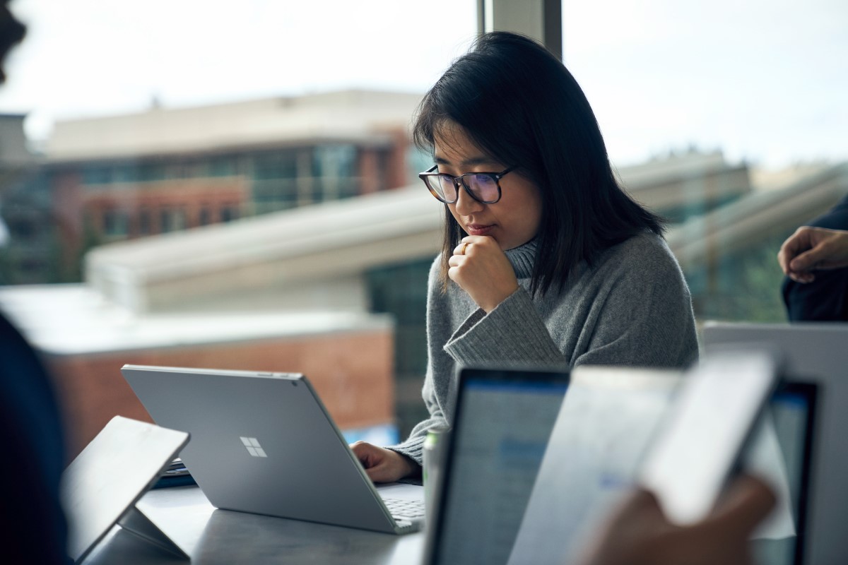 Woman with glasses sitting at a community table in common area reading on a Surface laptop. She looks as though she may be thinking, or learning