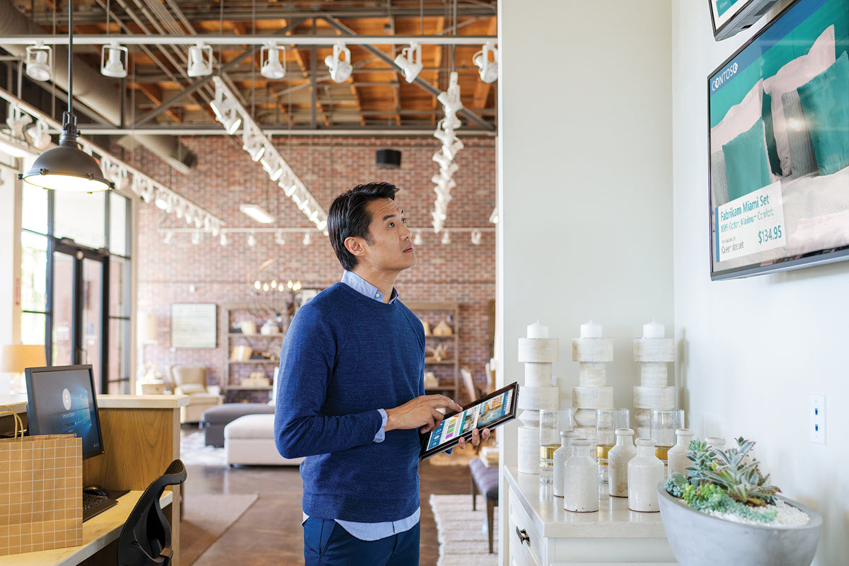 Male first line worker using laptop folded open as tablet, standing on home furnishings sales floor, looking at a wall-mounted customer support monitor which displays an advertisement for bedding. A desktop monitor sits on a desk behind him