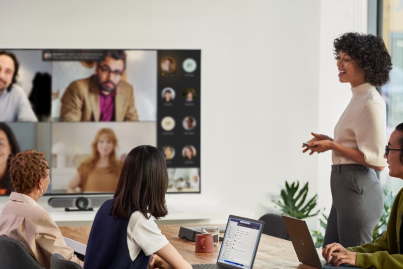A female speaker presenting in a Microsoft Teams Room meeting while people in the conference room are viewing remote attendees in Gallery Mode with participant chat on the front of room screen​