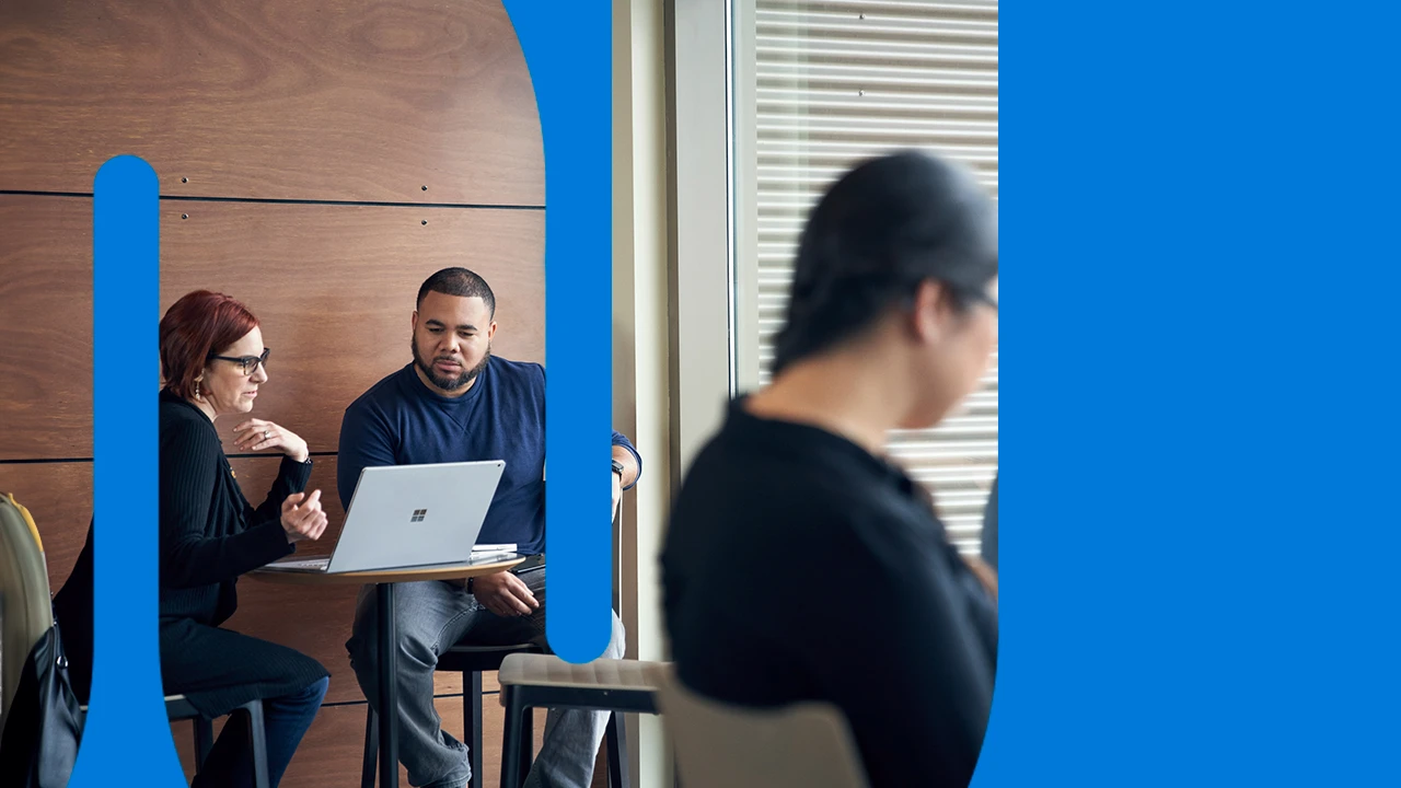 Man and woman meeting together and reviewing content on a Surface laptop at a high table with two blurred women in the foreground