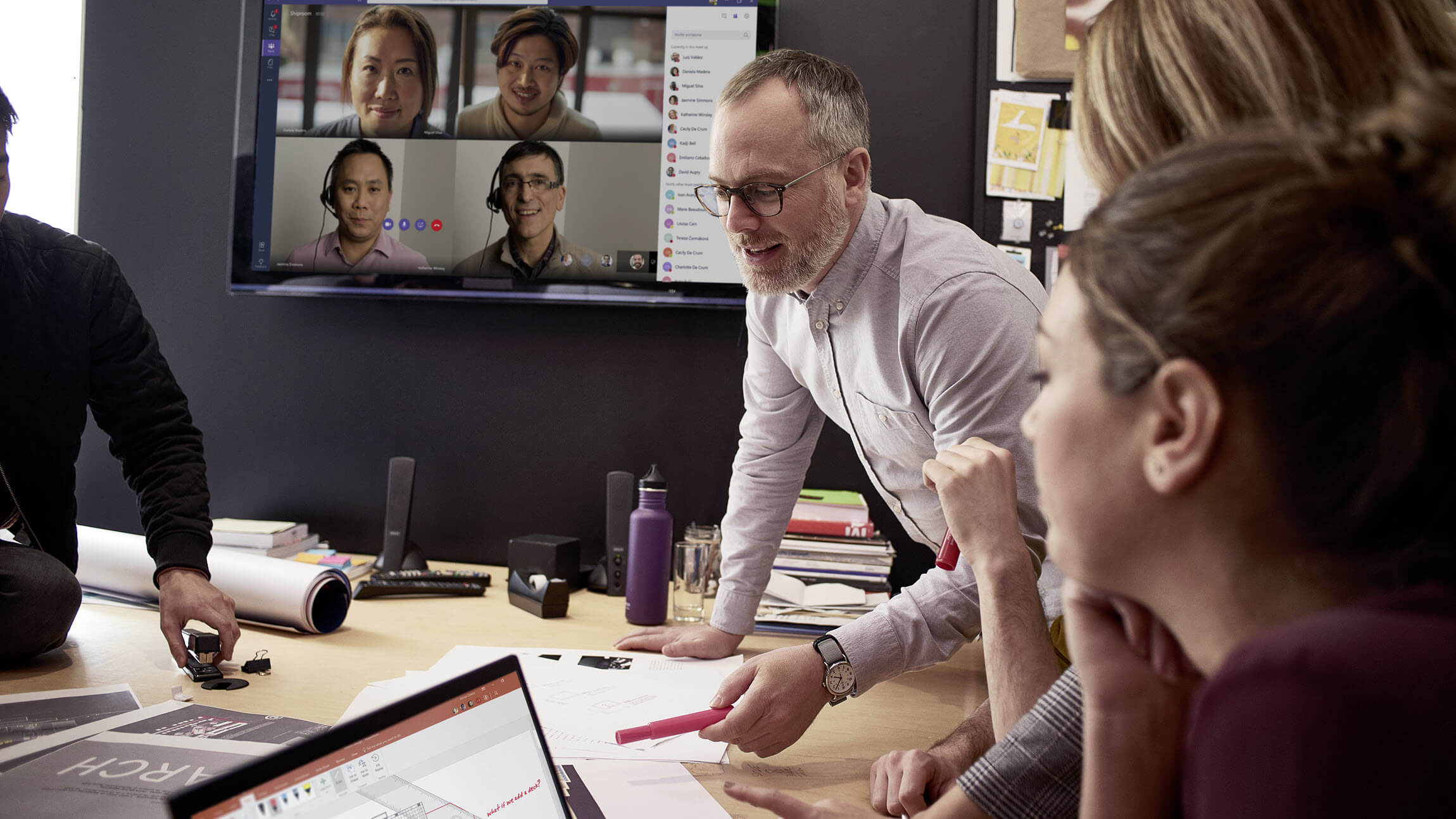 One person speaks at a conference room table as four people look on via a screen on Microsoft Teams and several others watch from in the room.